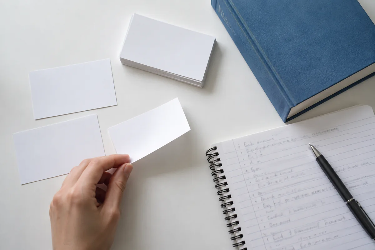 A learner holds a blank flashcard beside a notebook and a closed book.